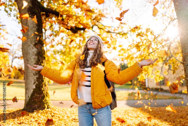 Obraz A sweet woman is having fun with fallen leaves in an autumn park. A beautiful woman tosses autumn leaves into the air at sunset. Concept of seasonality, fun, and relaxation.