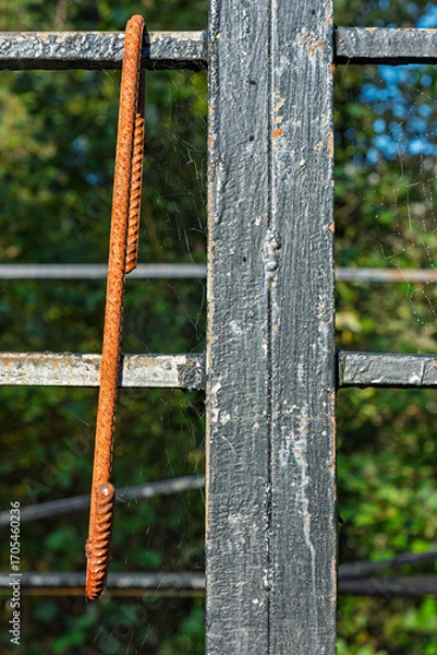 Obraz A rusty metal bar is suspended from a deteriorating structure, with vibrant green trees providing a natural backdrop under clear blue skies.