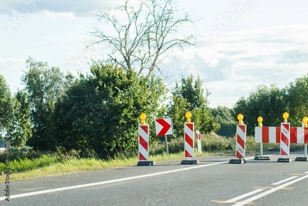 Fototapeta Road construction scene featuring traffic barriers and warning signs, surrounded by lush greenery and a clear sky, indicating ongoing roadwork and safety measures