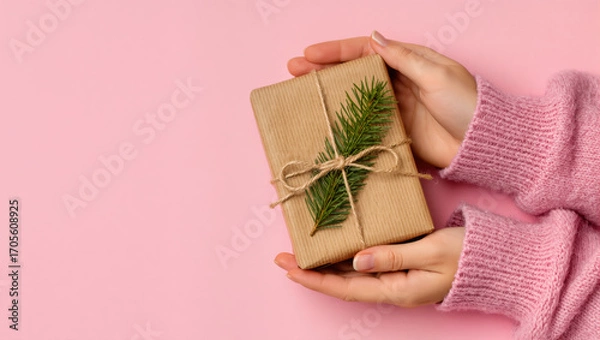 Fototapeta Christmas gift in eco wrapping with fir twig and twine held in female hands in pink sweater on pastel pink background
