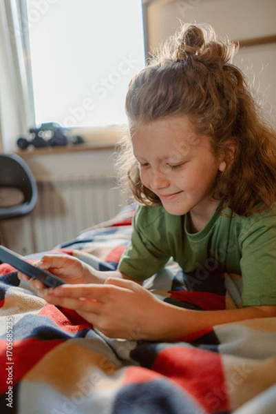 Fototapeta A boy relaxes on a colorful blanket while using his smartphone. He smiles as he engages with the device, showcasing a moment of leisure in a warm, inviting room during the day.