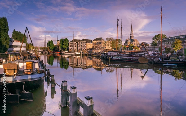 Fototapeta Golden hour at the historic harbor in Gouda, Netherlands, with old boats, calm water reflections, and a church in the background.