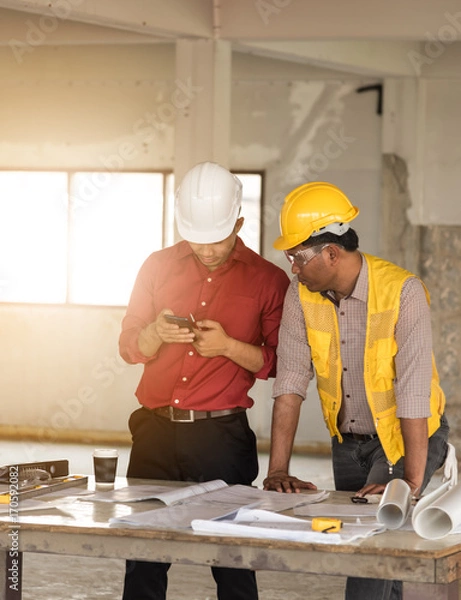Obraz Team of architects engineer working together Asian businessman with blueprint and Craftsman tool on table at construction site.using smart phone consult something.