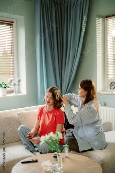 Fototapeta Two friends relax in a bright living room while one styles the other's hair. They sit comfortably on a sofa, surrounded by natural light and greenery, enjoying their time together.