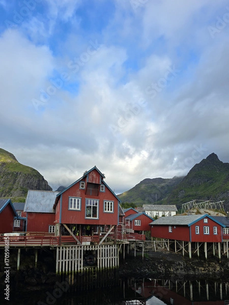 Fototapeta Colorful red cabins perched over the waterfront in A Lofoten, Norway, are framed by dramatic mountains and cloudy skies, creating a picturesque view that captures the essence of Norwegian coastal life