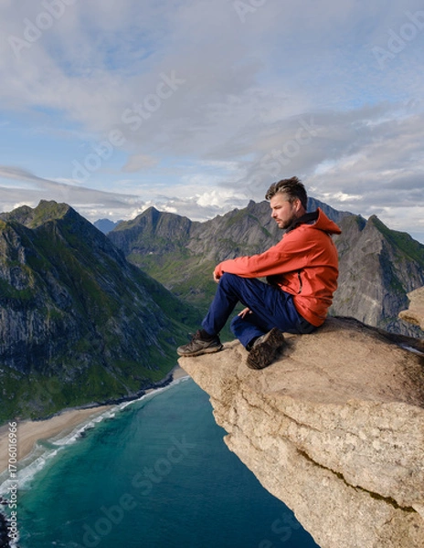 Fototapeta A hiker sits on the edge of a rocky precipice on the Ryten Kvalvika Beach Trail, admiring breathtaking views of the turquoise waters and rugged landscape of Lofoten, Norway.