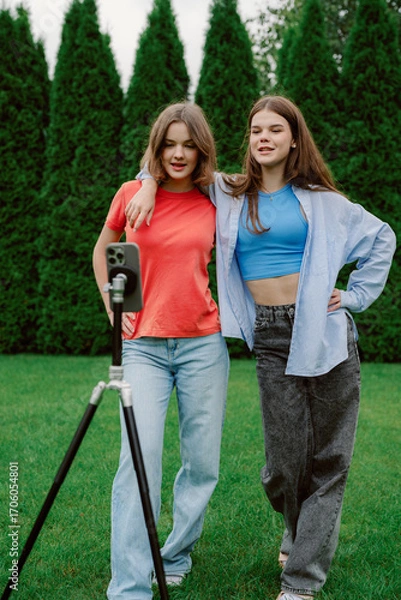 Fototapeta Two young women stand together in a park, smiling and posing for a video recording. Surrounded by green trees, they enjoy a playful moment in casual outfits.
