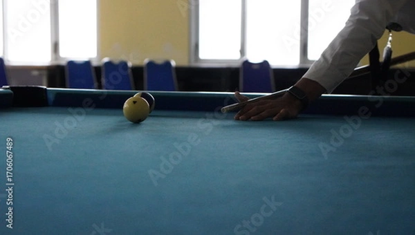 Fototapeta player’s arm extended over green pool table, hand positioned to guide cue ball near yellow-striped ball, indoor setting with chairs and windows softly lit in background