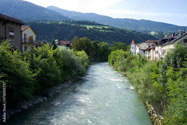 Fototapeta Serene river flowing through lush greenery and charming houses with mountains in the background, perfect for nature lovers