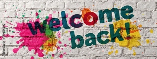 Fototapeta Welcome back!" text art on a white brick wall with colorful paint splashes, offering a bright, cheerful, & inviting message