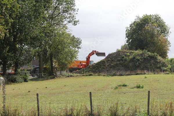 Fototapeta a crane is digging rubble and ground in a tipping trailer at a farm and a green meadow with trees in front in the countryside