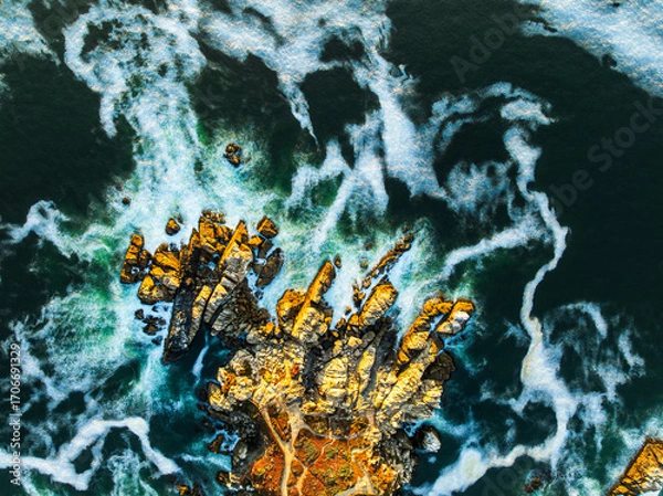 Obraz An abstract aerial long exposure of a rugged Pacific coastline, with golden-lit rocks surrounded by silky, white turbulent water at sunset