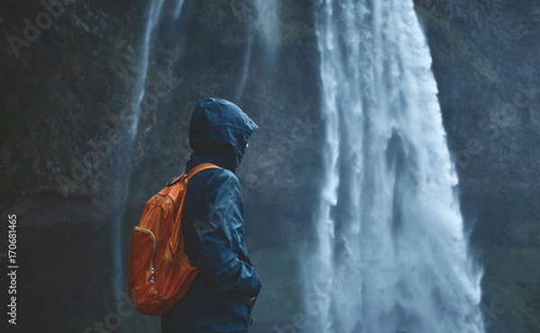 Obraz Girl in waterproof clothing stands under the Seljalandsfoss waterfall in Iceland. back view, woman with small orange backpack