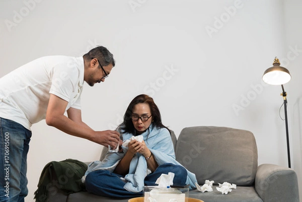 Fototapeta Man giving water and caring for sick woman with blanket while she sits on sofa suffering from cold at home