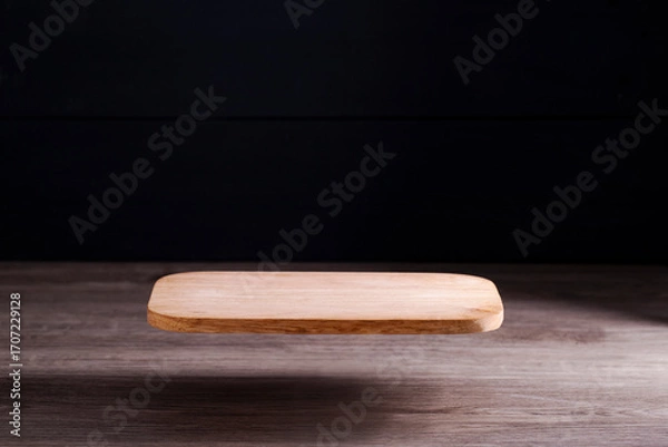 Fototapeta A brown wooden cutting board or board floats on the floor in an atmospheric light coming from the side, dark background, light reflecting on the floor.