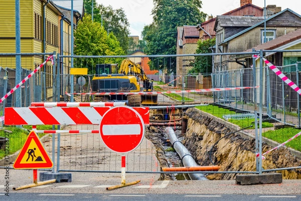 Fototapeta Street under construction with heavy machinery, excavation, and pipeline installation. Road closed with warning signs and safety barriers.