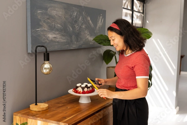 Fototapeta Woman decorating cake with fresh strawberries and cream in modern kitchen