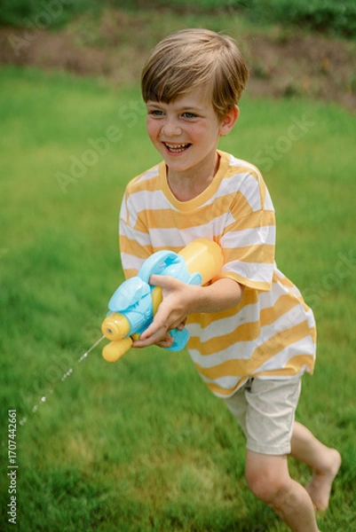Fototapeta A young boy wearing a striped shirt and shorts joyfully runs across a green lawn while aiming a colorful water gun. The bright summer day enhances his playful spirit.