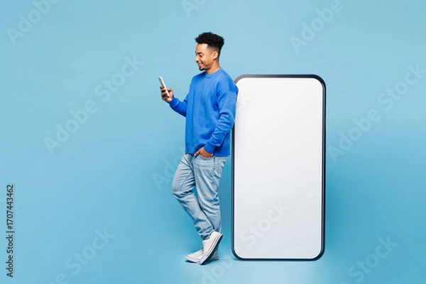 Fototapeta Full body side view young man of African American ethnicity in sweatshirt casual clothes stand near big huge blank screen mobile cell phone with area use smartphone isolated on plain blue background
