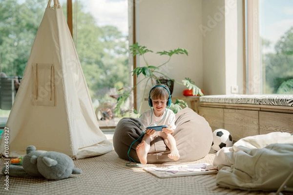 Fototapeta A young boy sits on a bean bag in a sunlit playroom, listening to a story on an electronic device while surrounded by toys, a tent, and bright green plants.
