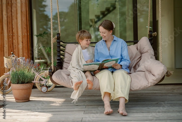 Fototapeta A mother reads a story to her child while sitting on a comfortable swing in their garden. It is a warm and sunny day, perfect for outdoor bonding time and imaginative adventures.