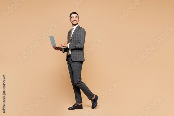 Fototapeta Full body side view young successful employee IT business man corporate lawyer wear grey suit shirt tie work in office hold use laptop pc computer look aside isolated on plain beige background studio.