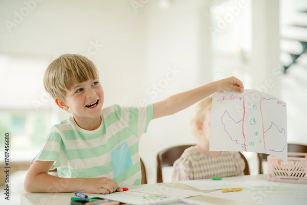 Fototapeta A young child proudly displays a colorful drawing while sitting at a table. The atmosphere is bright and cheerful, with another child engaged in activities nearby.