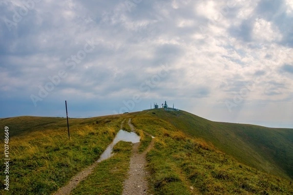Fototapeta Hiking trail leading to a mountain summit with a radar or communication station under dramatic clouds. Remote scenic landscape in the hills after rain.
