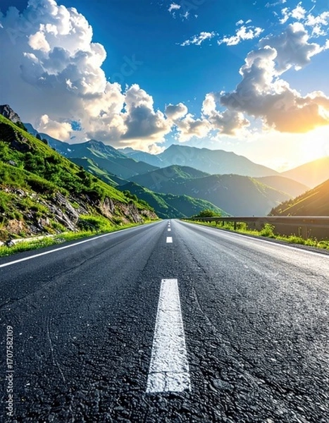 Fototapeta A long black asphalt road stretches towards distant green mountains, set against a bright blue sky with scattered puffy white clouds