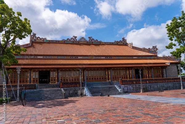 Fototapeta A traditional royal building with an orange tiled roof and detailed decorations stands on a vast brick courtyard in Hue, under a bright, cloudy sky.