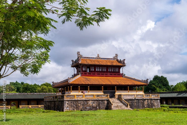 Fototapeta A classic two-story pavilion with an orange roof and stone base stands on a green lawn in Hue, framed by tree branches under a bright, cloudy sky.