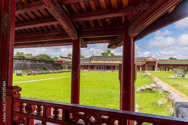 Fototapeta A peaceful courtyard in the Hue Imperial City, featuring a large green lawn, traditional buildings, and weathered stone walls, is viewed from under a red-pillared pavilion.