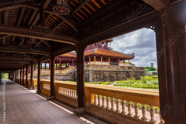 Obraz A tranquil garden courtyard with traditional buildings and manicured hedges is viewed through the dark wooden corridor of an ancient palace in Hue under a cloudy sky.