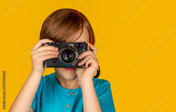 Fototapeta Boy with camera. Teen boy looks through a vintage camera and trying the photographer profession. Studio portrait over yellow background. Boy using a cameras