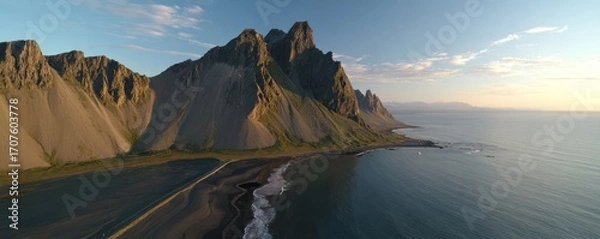 Fototapeta Aerial view of dramatic, rugged mountains meeting the ocean coastline under a bright sky with a sandy beach in the foreground