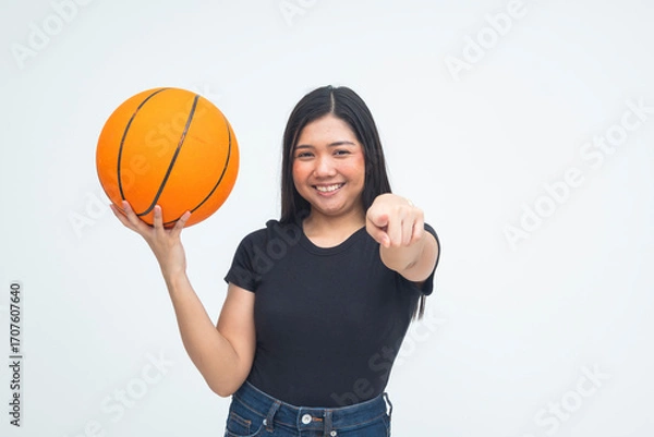 Fototapeta A bold young Asian woman in a half body shot smiling with pride while holding a basketball in one hand and pointing directly at the camera challengingly. Isolated on a white background.