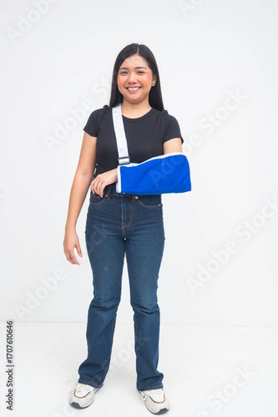 Fototapeta An optimistic young Asian woman in a full body shot standing with a bright smile while wearing an arm sling despite her injury. Isolated on a white background.
