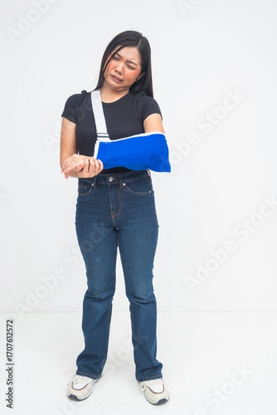 Fototapeta An injured young Asian woman in a full body shot standing with an arm sling, grimacing in pain and discomfort. Isolated on a white background.
