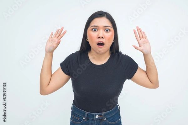 Fototapeta An astonished young Asian woman in a half body shot raising her hands with mouth open and wide eyes, expressing a wow and unbelievable reaction. Isolated on a white background.