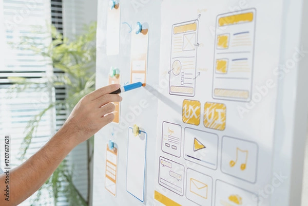 Fototapeta A young designer is working on a UX UI project, analyzing mobile app wireframes and choosing color palettes on a whiteboard. He focuses on brainstorming ideas to improve user experience.