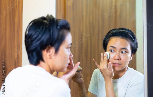 Fototapeta Young Asian woman gently cleanses her face with a cotton pad and cleanser in bedroom. Skincare routine focuses on makeup removal, self care, and achieving a clean, healthy face with mirror reflection.