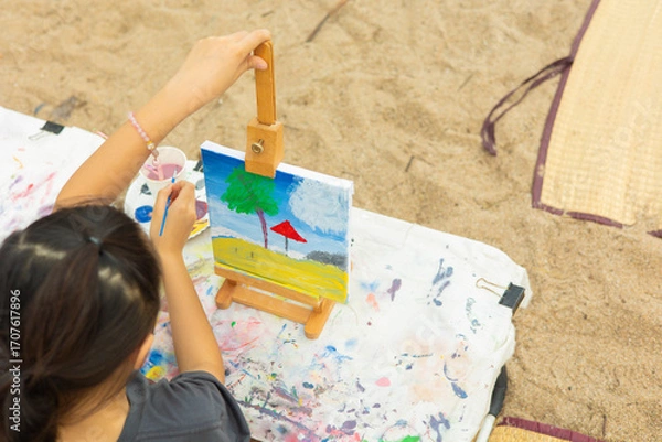 Fototapeta Close-up side view of a young girl focused on her painting at an easel outdoors. This shows the intense concentration and creativity of a child engaged in an artistic hobby or art class.