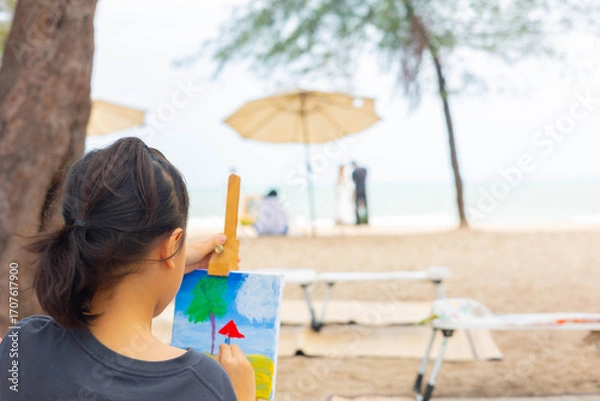 Fototapeta Over-the-shoulder view with blurred background of a young girl painting a seascape on an easel at beautiful beach, representing childhood creativity, outdoor learning, and a peaceful artist hobby.