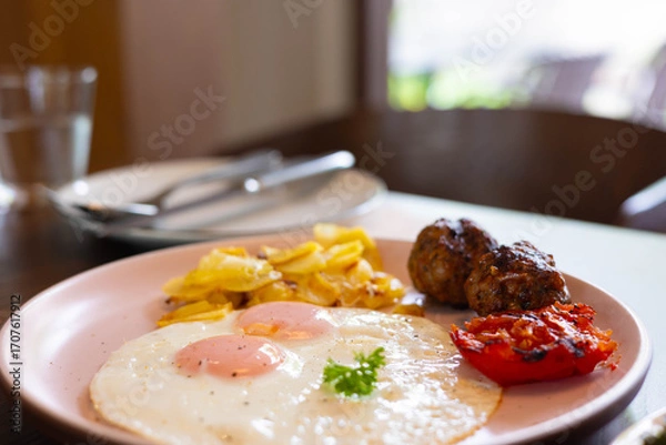 Fototapeta Selective focus close-up of breakfast plate with fried eggs, potato wedges, grilled tomato, and meatballs in morning. Food photography, restaurant menu design, healthy brunch concepts.