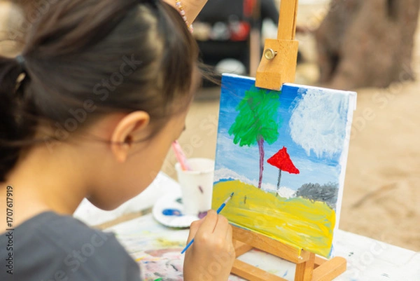 Fototapeta Close-up and selective focus of young girl painting a palm tree and sea with beautiful color on canvas. This shows creativity of a child's artistic hobby in a natural, outdoor beach setting.
