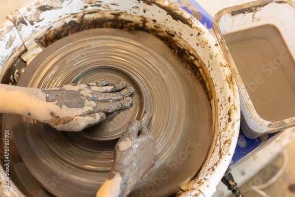 Fototapeta Top view of female hands shaping clay on pottery wheel in selective focus. Artistic handmade ceramic, learning pottery craft, creative process, traditional artisan skill, hobby and craftsmanship.