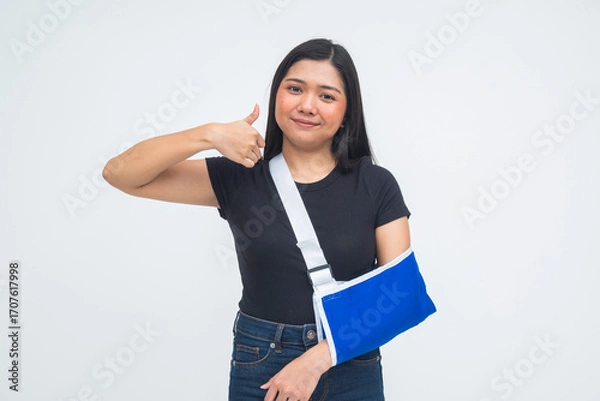 Fototapeta A hopeful young Asian woman in a half body shot wearing an arm sling and giving a thumbs up with a smile, demonstrating optimism and a bright outlook. Isolated on a white background.