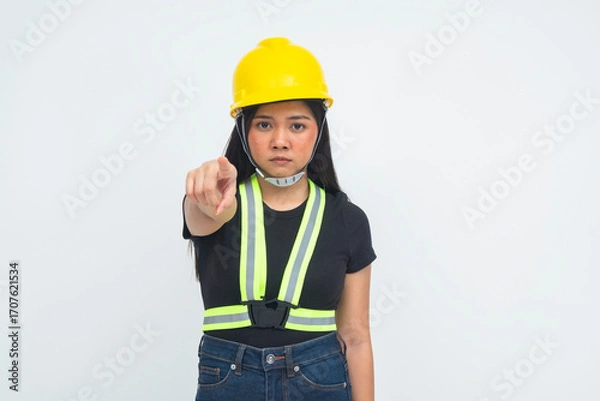 Fototapeta A strict young female engineer in a half body shot wearing a protective helmet, pointing her finger at the camera with a scolding and confrontational expression. Isolated on a white background.