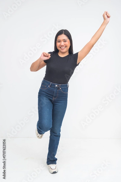 Fototapeta An enthusiastic young Asian woman in a full body shot almost jumping with one foot up, fist raised, and smiling excitedly. Isolated on a white background.