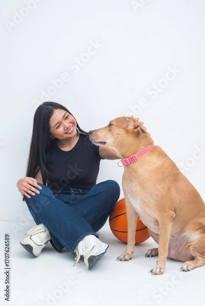 Fototapeta A cheerful young Asian woman captured sitting beside a basketball on the floor, smiling and gently petting a dog in a friendly manner. Isolated on a white background.